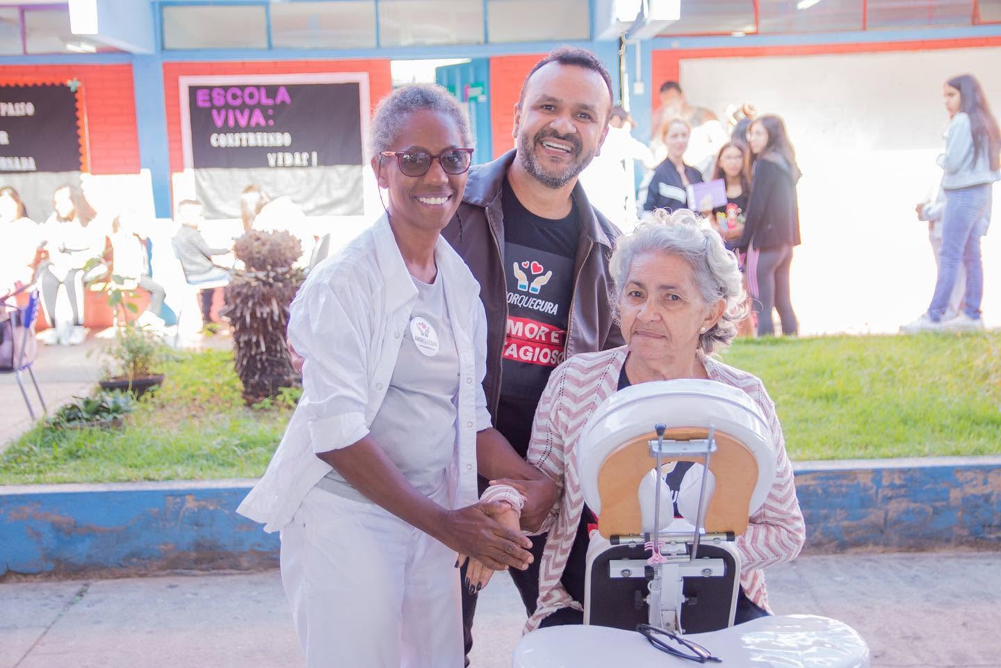 Uma pessoa em cadeira de rodas está em primeiro plano, ao lado de duas pessoas; uma delas usa camiseta com a marca Amor que Cura.