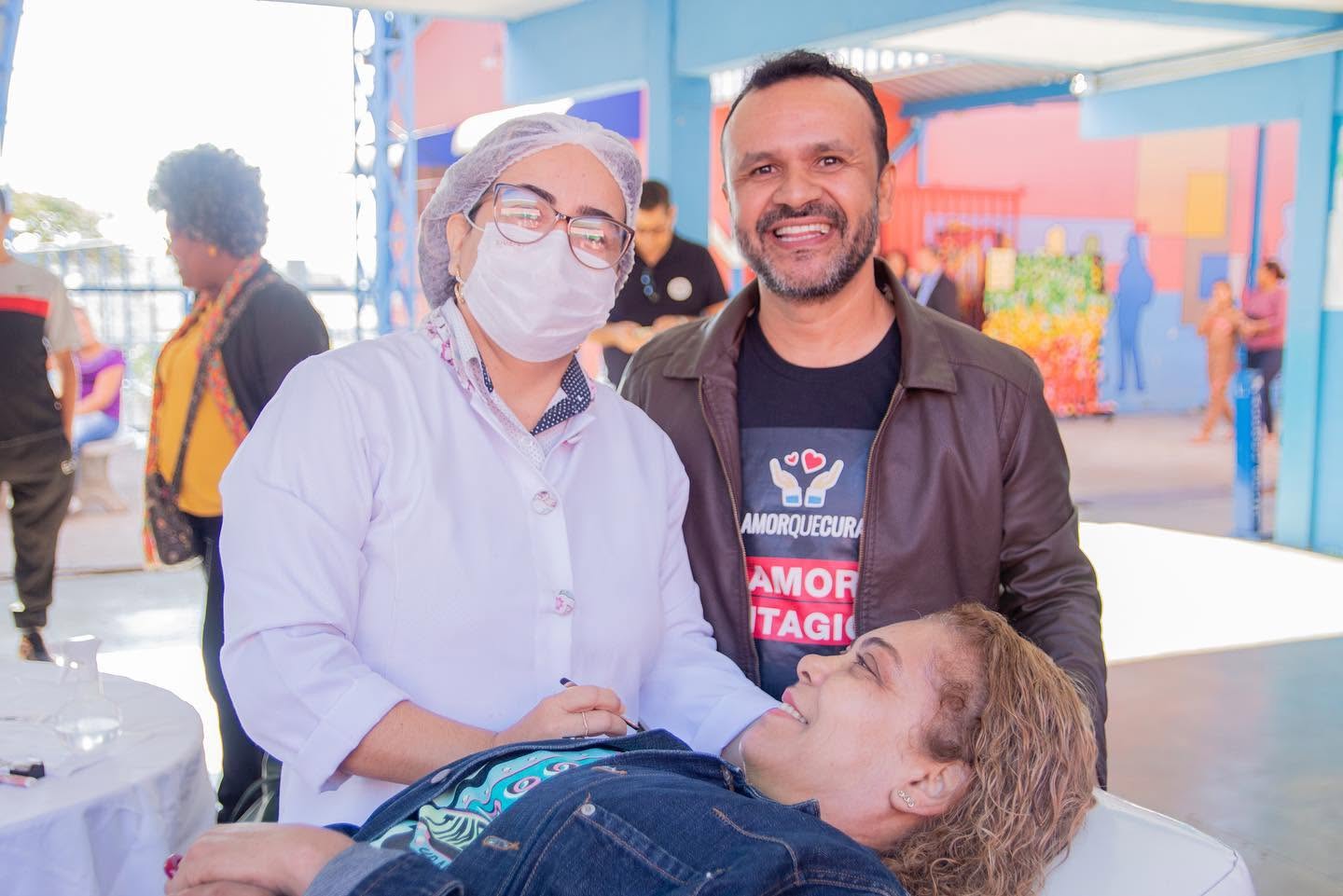 Uma pessoa com roupa branca e máscara está ao lado de uma pessoa deitada; ao fundo, outra pessoa usa camiseta com a marca Amor que Cura.