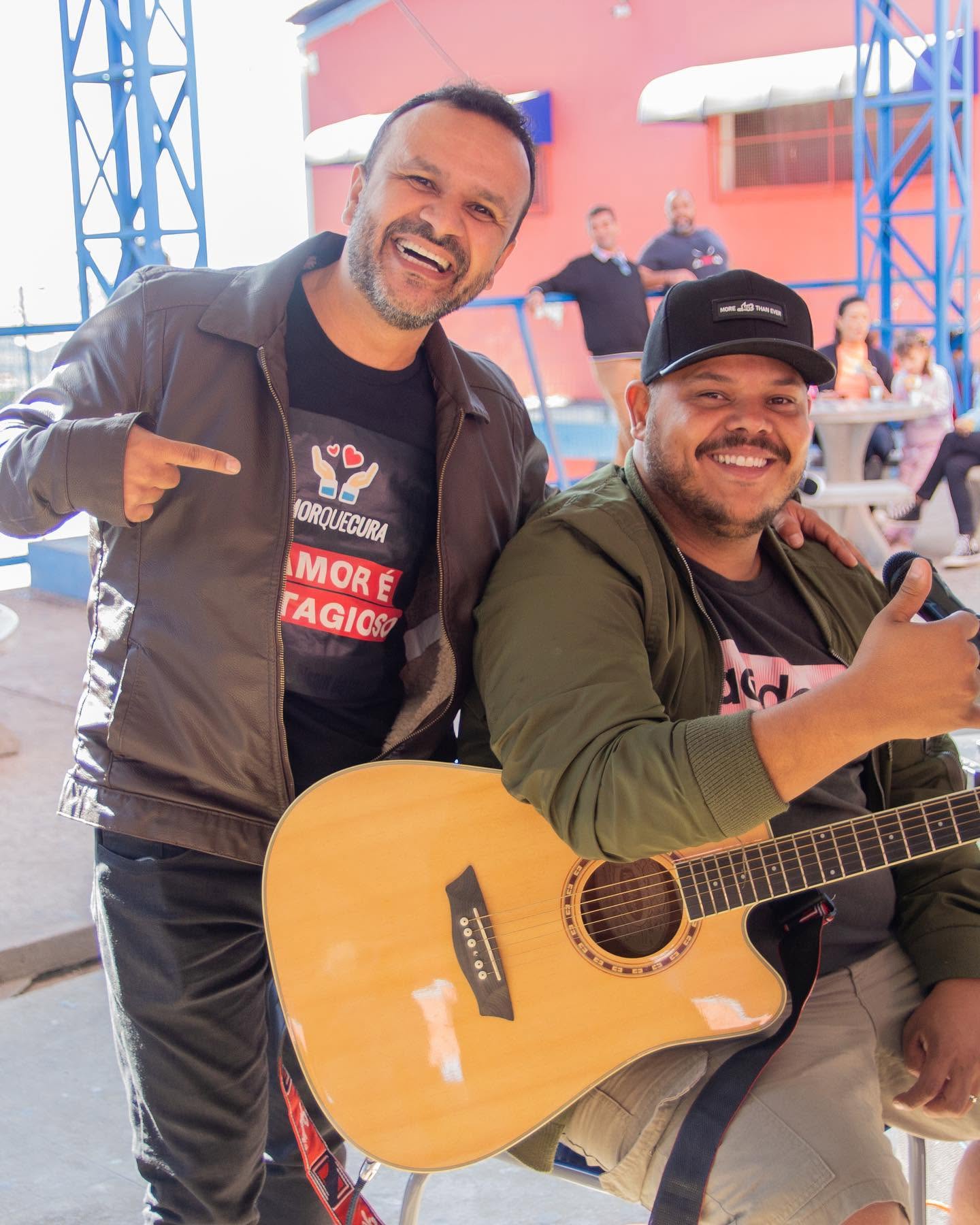 Duas pessoas reunidas; uma delas segura um violão e veste camiseta com a marca Amor que Cura.